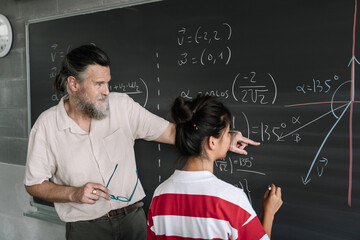 Young Female Asian Teenager Student doing Maths Exercises on Blackboard with help of the teacher in...