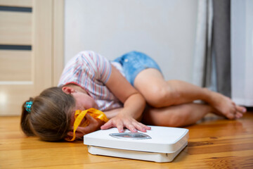 Distressed girl lying on the floor next to a weighing scale and measuring tape