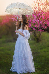girl bride in vintage dress with white lace umbrella in park near blossoming tree with pink flowers