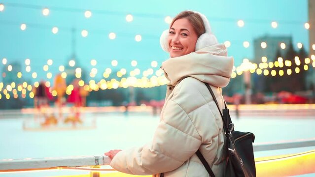 Woman in earmuff and warm clothes smiling at camera while standing outside ice rink. Beautiful young lady enjoying Christmas holidays in city with blurred Christmas lights in background.