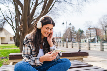 One young girl is listening to music on her wireless headphones and using her phone outdoors on a sunny day	