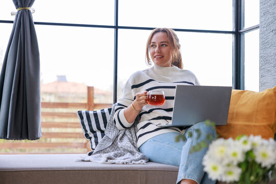 Young woman using a laptop computer while relaxing on the windowsill sofa at home