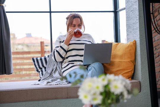 Young woman using a laptop computer while relaxing on the windowsill sofa at home