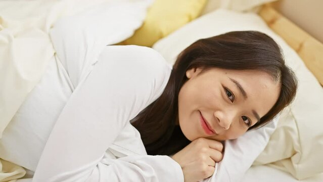 A relaxed asian woman lying in bed, enjoying leisure time in a cozy bedroom setting.