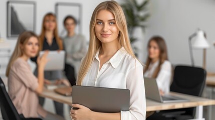 Obraz premium Portrait of attractive female entrepreneur in casual clothes holding laptop in hand standing in office, looking at camera. Startup business team discussing project sitting at desk on background.