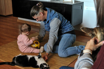 Caucasian father plays with his baby and his cat on the living room floor while mother and her...