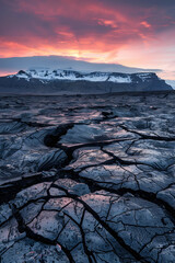 Iceland, sunset, cracked black lava floor with snowcapped mountains in the background - obrazy, fototapety, plakaty