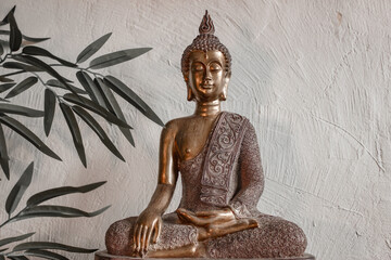 Estatua de Buda de bronce en fondo blanco.
La estatua está ubicada contra un fondo blanco texturizado y está rodeada de hojas de bambú. La composición transmite una sensación de paz y espiritualidad.
