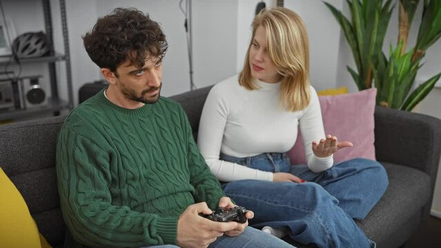 Couple in a living room, man holding a game controller and woman gesturing with a puzzled expression, showcasing a modern apartment interior.