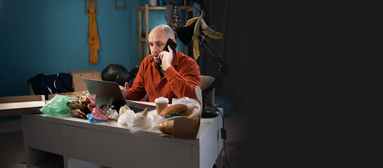 Man working in Messy, cluttered room. Bearded freelancer making phone call and using laptop at home