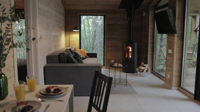 Cozy living room in cabin house, chimney fire place with fire burning at the living room in tiny house, wooden wall and large window on the background, warm light from floor lamp, slow motion.
