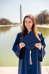 Young woman graduate holding a cap, smiling with a reflecting pond and Washington Monument in the background.