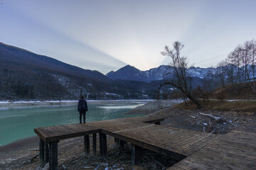 Winter sunset behind a mountain peak of a winter alpine landscape with hiker on wooden pier at...
