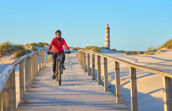 happy active senior woman cycling during moody golden hour at the beach of the atlantic coast below Barra Lighthouse near Aveiro, Portugal Aveiro, Portugal