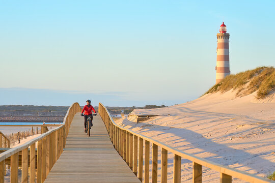 happy active senior woman cycling during moody golden hour at the beach of the atlantic coast below Barra Lighthouse near Aveiro, Portugal Aveiro, Portugal