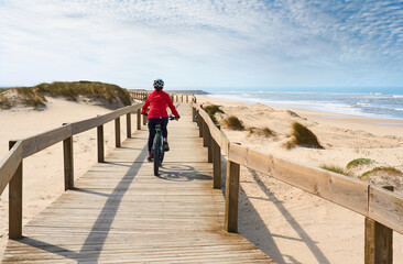 happy active senior woman cycling during moody golden hour at the beach of the atlantic coast of Aveiro, Portugal - obrazy, fototapety, plakaty