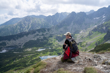Fototapeta premium Woman with backpack, hiking in the mountains, sitting and looks at the valley from the High Tatras. Sunny summer day with rain clouds in the distance.