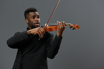 Elegant African American man in black suit playing violin with passion on gray background