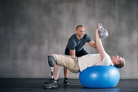 Trainer, man with a disability and prosthetic leg and dumbbell in physiotherapy, studio and gym ball. Male people, physiotherapy and amputee for wellness, fitness and exercise in rehab center - Powered by Adobe