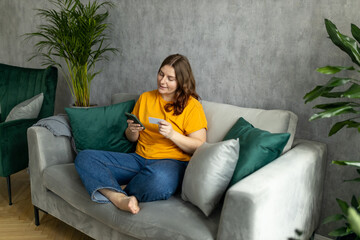 Obraz premium Cheerful young woman in casual clothes sit on couch spending time in living room at home. Rest relax good mood leisure lifestyle concept. 30s female Using phone, hold credit bank card, full body
