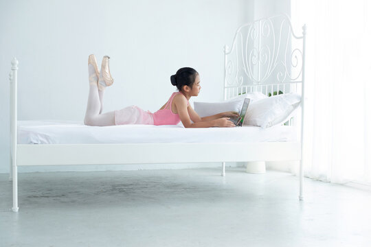 Little ballerina girl lies looking at laptop screen on bed in white bedroom