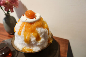 traditional Japanese sobert cold shaved ice dessert kakigori bingsu topped with orange marmalade sauce and fruit, oolong tea sauce on a black plate with white background and wooden table in summer - obrazy, fototapety, plakaty