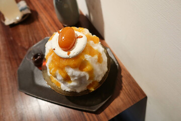 traditional Japanese sobert cold shaved ice dessert kakigori bingsu topped with orange marmalade sauce and fruit, oolong tea sauce on a black plate with white background and wooden table in summer