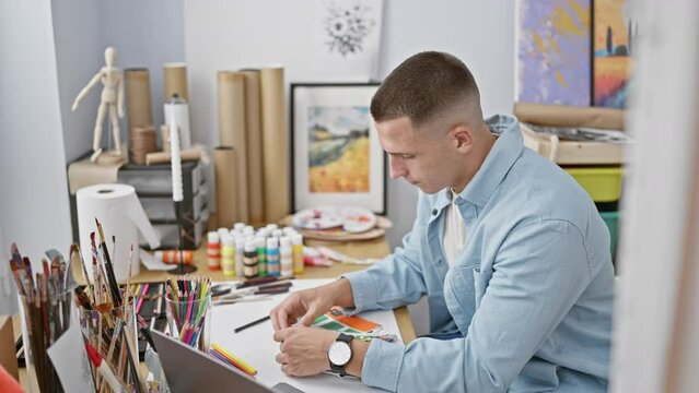 A focused man selecting colors from a palette in a studio filled with art supplies and paintings