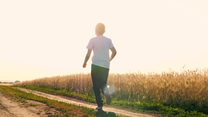 Cheerful teen boy running on dirt road at sunny sunset sunrise wheat field enjoy freedom happy...