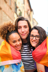 Three lesbian and trans women are hugging each other wrapped in a rainbow flag. Scene is joyful and celebratory.