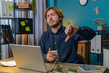 Dislike. Upset bored Caucasian businessman using smartphone at home office showing thumbs down sign...