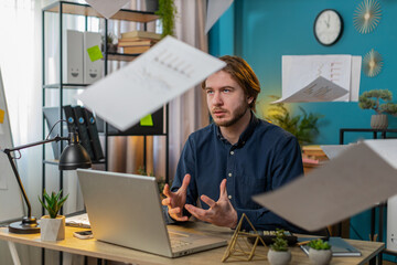 Frustrated upset young businessman works at home office desk looking at bills paperwork reports...