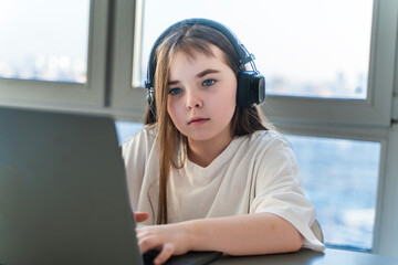Cute teenage girl with long hair in headphones sitting at laptop on window background. High quality...