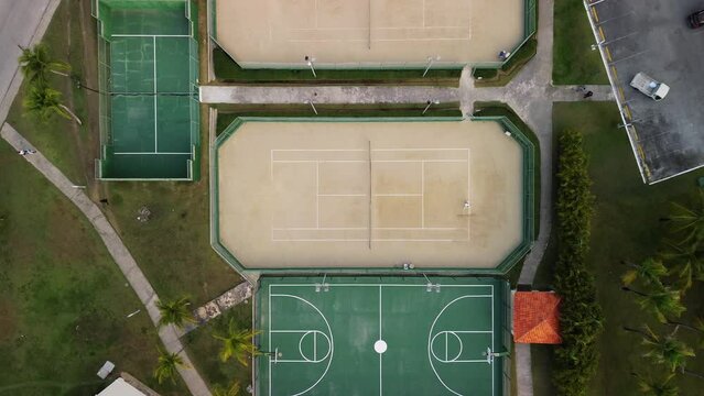 Person is seen from aerial top perspective, cleaning beige tennis court within sports facility that also features basketball. Camera smoothly zooms in on scene.