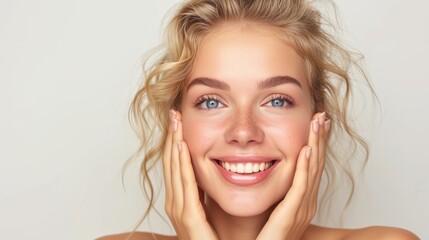 Fototapeta premium A young European blonde woman with a joyful expression against a white background. Woman with natural makeup, showcasing radiant skin and a bright smile. Curly hair frames her face, hands on cheeks