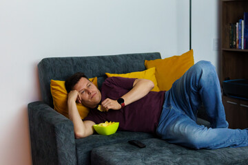 a man lying down is falling asleep on the sofa at home watching television