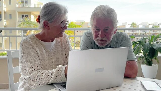 Cheerful senior couple using laptop enjoying technology, elderly 70 years old caucasian people sitting outdoors on terrace browsing, shopping on line, follow social media together