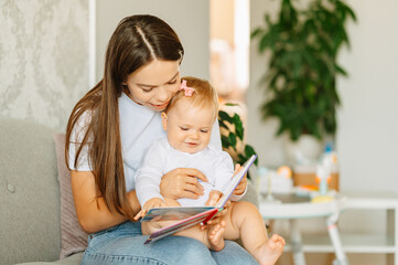 Young mother is holding her sweet daughter and reading a kids book to her at home.