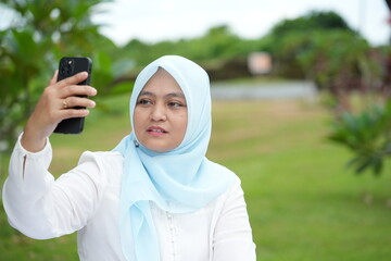 Asian woman in blue hijab is holding a cell phone with her facial expression while outdoors in the...