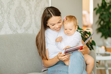 Young mother is holding her sweet daughter and reading a kids book to her at home.