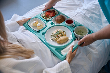 Nursing assistant serving meal to recumbent patient - obrazy, fototapety, plakaty