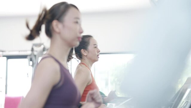 Two women are running on a treadmill. One of them is wearing a purple top. The other woman is wearing an orange top