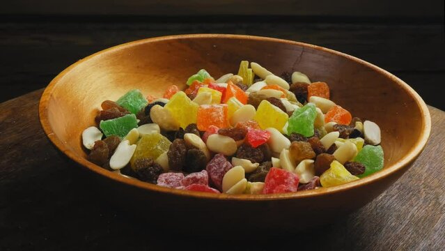 Mix of multi-colored dried fruits, candied fruits and scattered nuts in a wooden plate on a black background, rotating in a circle. Medium plan