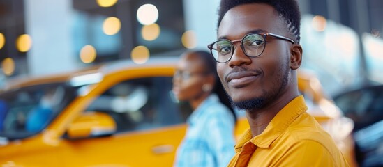 A man with glasses and a yellow shirt is standing in front of a yellow car - obrazy, fototapety, plakaty