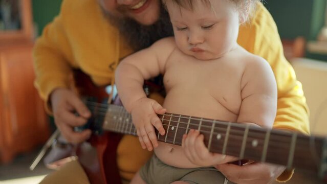 Smiling young father holds his little cute baby-boy on his knees and tries to teach her to play the guitar, cheerful toddler claps the guitar as if she playing it. Modern family, happy memories. Slow