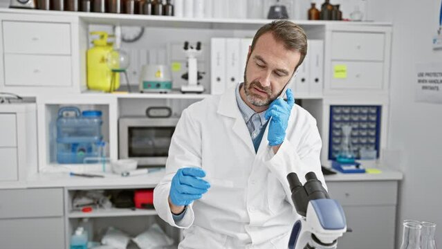 A bearded male scientist in a laboratory examines a slide while talking on the phone indoors.