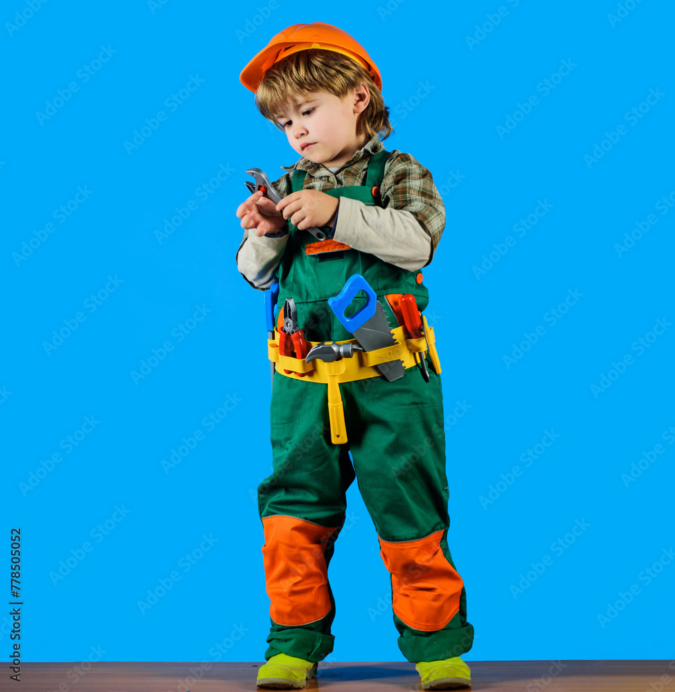 Wall mural Little builder in helmet and tool belt with adjustable wrench. Kid repairman in protective hard hat and overalls with tool belt of toy tools for repair. Child boy with set of toy construction tools.