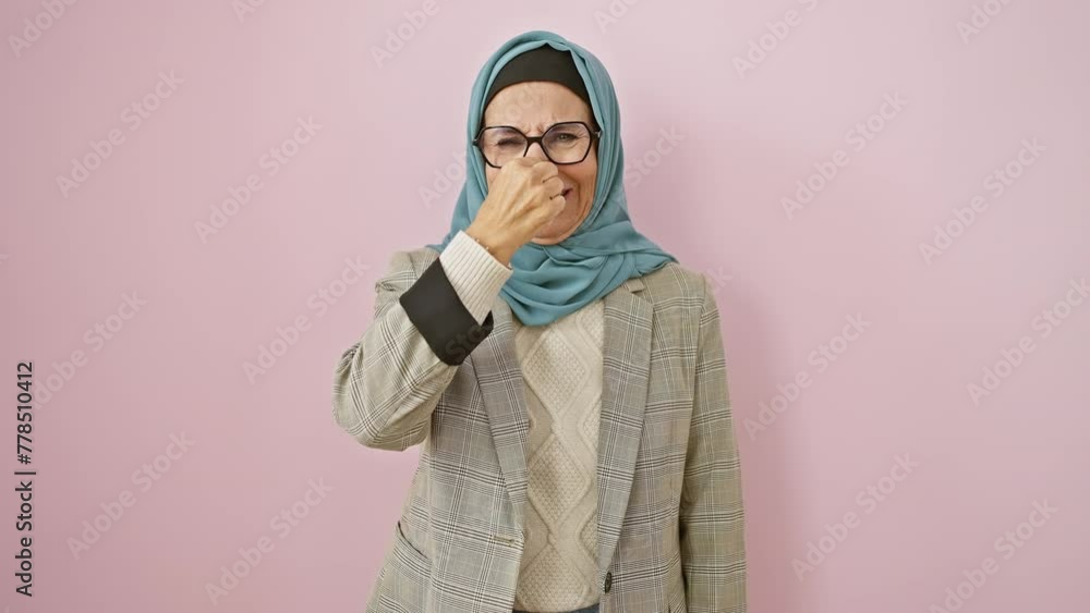 Sticker Middle-aged hispanic woman in hijab disgusted by intolerable stink, holding nose over stinky smell, isolated on pink background