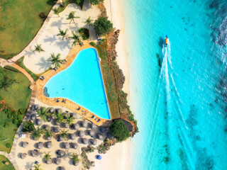 Aerial view of pool, white sandy beach with palms, umbrellas, swimming people, boat, blue ocean, at sunset. Summer vacation in Nungwi, Zanzibar island. Tropical landscape. Clear azure sea. Top view