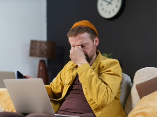 A man in a yellow jacket is sitting on a couch with his head in his hands. He is looking at a laptop and he is in a state of distress